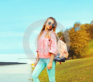 Portrait of stylish young blonde woman posing wearing pink knitted sweater and backpack outdoors in city park