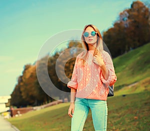 Portrait of stylish young blonde woman posing wearing pink knitted sweater and backpack outdoors in city park