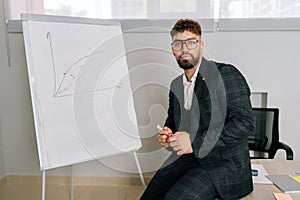 Portrait of stylish young businessman in glasses and suit drawing financial plan on flipchart and explaining to team of