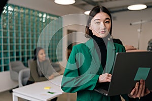 Portrait of stylish young businesswoman holding laptop in hand standing in office confident looking at camera. Diverse