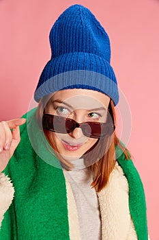 Portrait of stylish young girl in sunglasses, blue hat, green scarf and fur coat posing over pink background. Flirty