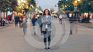 Portrait of stylish young lady tired of usual haste standing in the street among whizzing people and looking at camera