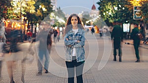 Portrait of stylish young lady tired of usual haste standing in the street among whizzing people and looking at camera