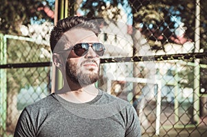Portrait of a stylish young man with a beard and black glasses on the sports field, metal grille