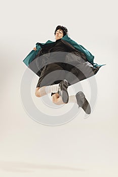 Portrait of stylish young man in black outfit and green coat posing, jumping  over grey studio background