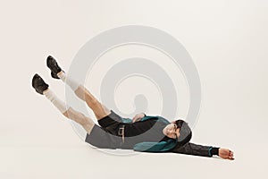 Portrait of stylish young man in black outfit and green coat posing, lying on floor  over grey studio background