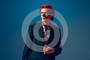 Portrait of stylish young man in official jasket and black glasses, posing, looking away  over blue studio