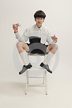 Portrait of stylish young man with piercing sitting on chair, drinking coffee  over grey studio background