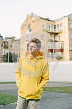 Portrait of stylish young man in yellow hoodie posing at camera on background of velodrome and old buildings. Vertical