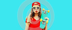 Portrait of stylish young woman blowing her lips with red lipstick holding banana and skateboard isolated on blue background