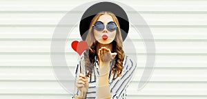 Portrait of stylish young woman blowing her red lips sending air kiss with red heart shaped lollipop wearing black round hat,