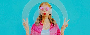 Portrait of stylish young woman blowing lips in wireless headphones listening to music wearing pink jacket, sunglasses over blue