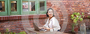 Portrait of stylish young woman, brunette girl with laptop, sitting outdoors and using computer