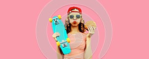 Portrait of stylish young woman with burger fast food and skateboard wearing baseball cap, sunglasses on pink background