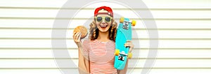 Portrait of stylish young woman with burger fast food and skateboard wearing baseball cap, sunglasses on white background