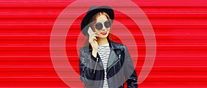 Portrait of stylish young woman calling on smartphone wearing black round hat, leather biker rock jacket on red background