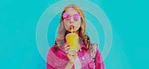 Portrait of stylish young woman drinking juice wearing pink jacket, sunglasses over blue background