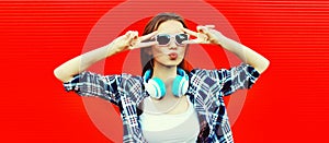 Portrait of stylish young woman with headphones listening to music blowing her lips sends air kiss on red background