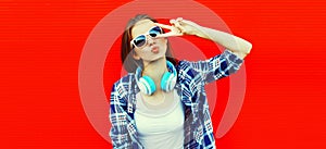 Portrait of stylish young woman with headphones listening to music blowing her lips sends air kiss on red background