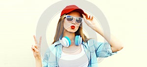 Portrait of stylish young woman with headphones listening to music blowing her lips sends air kiss wearing red baseball cap on