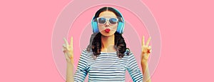 Portrait of stylish young woman in headphones listening to music blowing her lips sends sweet air kiss on pink background