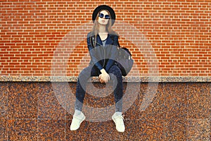 Portrait of stylish young woman model wearing black round hat, leather jacket in rock style on city street on brick wall