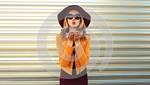 Portrait of stylish young woman posing blowing her lips sends sweet air kiss wearing sunglasses, round hat and jacket on gray