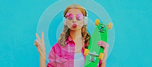 Portrait of stylish young woman with skateboard listening to music in wireless headphones wearing a pink jacket, sunglasses