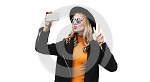 Portrait of stylish young woman taking selfie with smartphone blowing her lips wearing black coat, round hat isolated on white