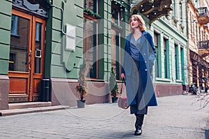 Portrait of stylish young woman wearing in blue coat walking holding purse outdoors. Spring fashion female accessories