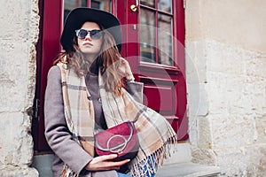 Portrait of stylish young woman wearing hat coat holding purse outdoors. Spring fashion female retro accessories outfit