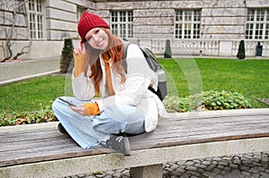 Portrait of stylish young woman, 25 years, sits on bench in park and uses mobile phone, reads online news, messages or
