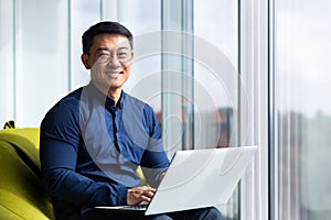 Portrait of successful asian businessman inside office, man smiling and looking at camera, using laptop at work near