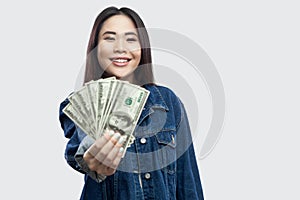 Portrait of successful attractive young asian girl in blue denim jacket standing, holding many dollars, demonstration and looking