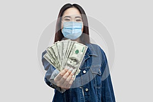 Portrait of successful attractive young asian woman with medical mask standing, holding dollars, demonstration and looking at