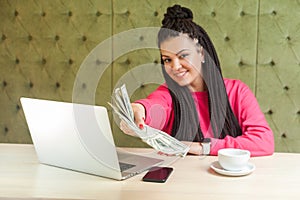 Portrait of successful attractive young woman with black dreadlocks in pink blouse is sitting, holding many dollars, giving you
