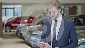 Portrait of a successful bearded seller in glasses dressed in a business suit standing in the hall of a modern car