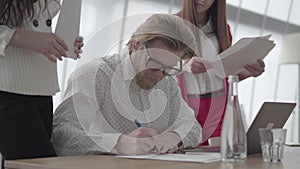 Portrait successful blond man in glasses sitting in a light comfortable office with netbook at the table signing