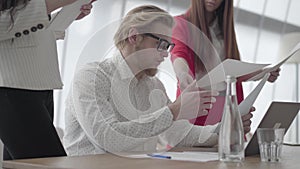 Portrait successful blond man in glasses sitting in a light comfortable office with netbook at the table signing