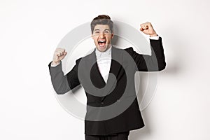 Portrait of successful businessman in black suit, become champion, raising hands up and shouting yes, triumphing and