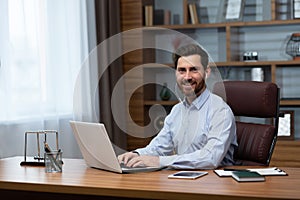 Portrait of successful businessman investor inside office, mature man smiling and looking at camera using laptop at work