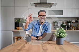 Portrait of successful businessman showing OK sign while working over laptop on desk at home
