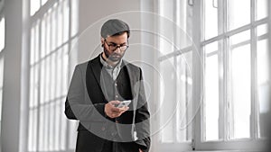 Portrait of successful businessman using modern smartphone in building with white walls, medium shot