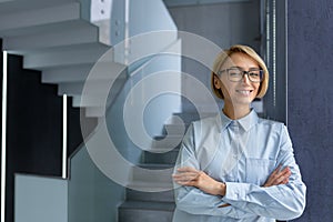 Portrait of successful businesswoman inside modern office building, female worker with crossed arms smiling and looking