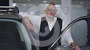 Portrait of successful confident Caucasian businessman in eyeglasses with mohawk haircut posing at open luxurious car in