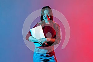 Portrait of successful confident office worker, beautiful girl in casual pullover standing, holding closed laptop