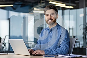 Portrait of a successful and confident young man in a blue shirt sitting in the office at a desk, working on a laptop