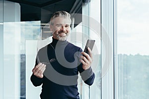 Portrait of a successful gray-haired man, senior businessman boss in the office smiling and looking at the camera by the window,