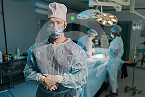 Portrait of successful male doctor standing posing looking at camera with crossed arms in operating theater with dark