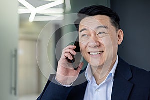 Portrait of successful mature asian businessman inside office at workplace, senior boss talking on phone, smiling, man